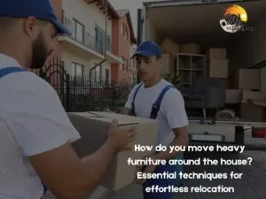 Our Blog 5 B&W Movers Two movers in blue hats and white shirts expertly load a cardboard box into a moving truck parked near a residential building. A logo and text about relocation techniques are visible, showcasing their skill in handling heavy furniture.